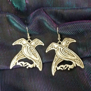 May include: A pair of silver earrings featuring a detailed design of two crows facing each other. The earrings are on a black fabric background.