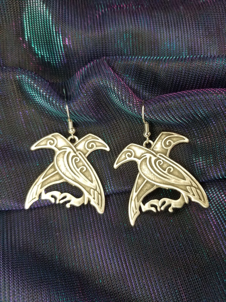 May include: A pair of silver earrings featuring a detailed design of two crows facing each other. The earrings are on a black fabric background.