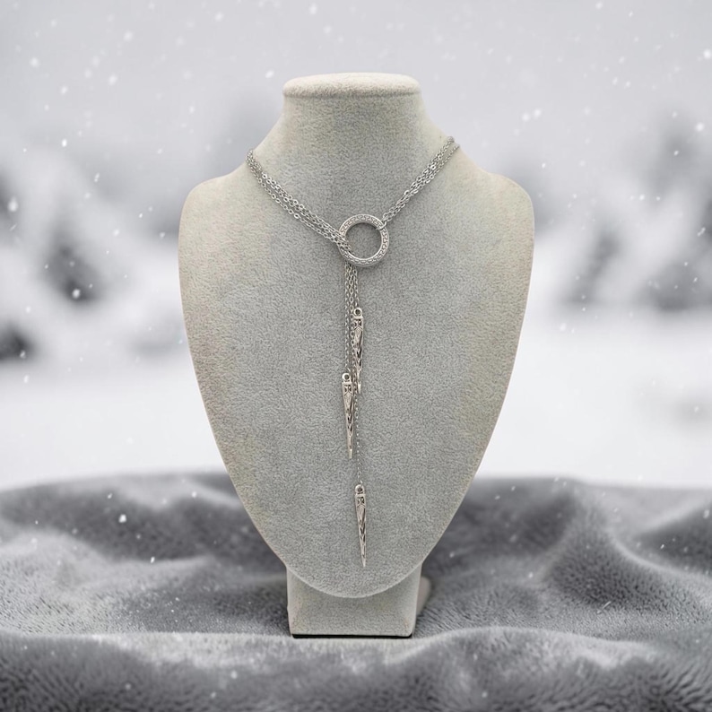 May include: A silver-toned lariat necklace featuring a central circular ring with a textured surface. Three delicate chains descend from the ring, each ending in a slender, pointed spike pendant. The necklace is displayed on a neutral-colored bust.