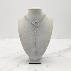 May include: A silver-toned lariat necklace featuring a central circular ring connector. Three delicate chains of varying lengths descend from the ring, each ending in a slender, pointed metallic spike charm with a textured, etched surface finish.