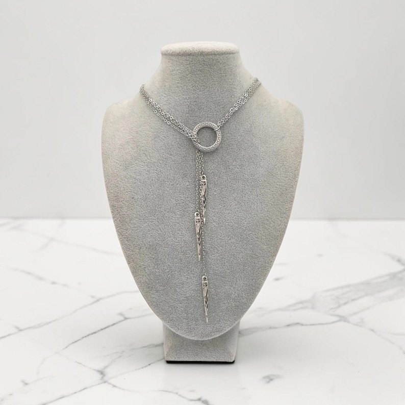 May include: A silver-toned lariat necklace featuring a central circular ring connector. Three delicate chains of varying lengths descend from the ring, each ending in a slender, pointed metallic spike charm with a textured, etched surface finish.
