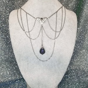 May include: A silver chain necklace with a delicate floral design and a dangling purple crystal. The necklace has multiple layers of chains and is displayed on a white mannequin.