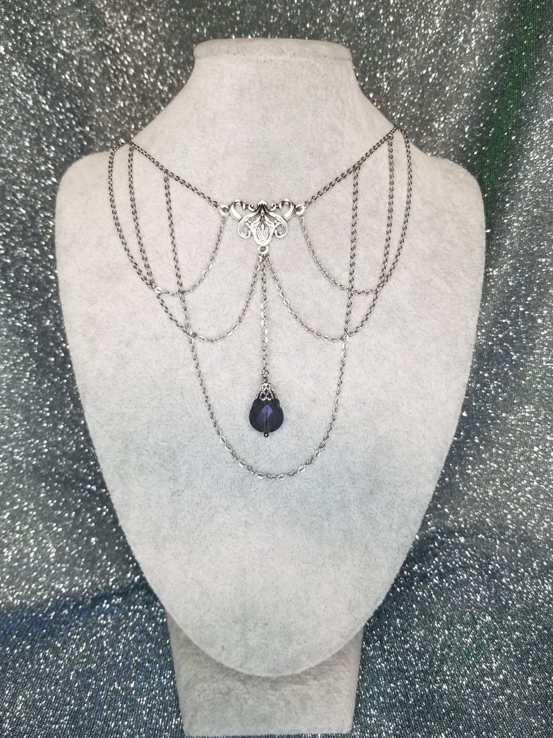 May include: A silver chain necklace with a delicate floral design and a dangling purple crystal. The necklace has multiple layers of chains and is displayed on a white mannequin.