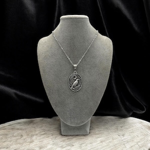 Puede incluir: Un collar con colgante ovalado de tono plateado que presenta un dise&ntilde;o en relieve de un cuervo posado sobre una rama bajo una luna creciente. El colgante cuelga de una delicada cadena plateada, mostrando detalles intrincados y un acabado met&aacute;lico.