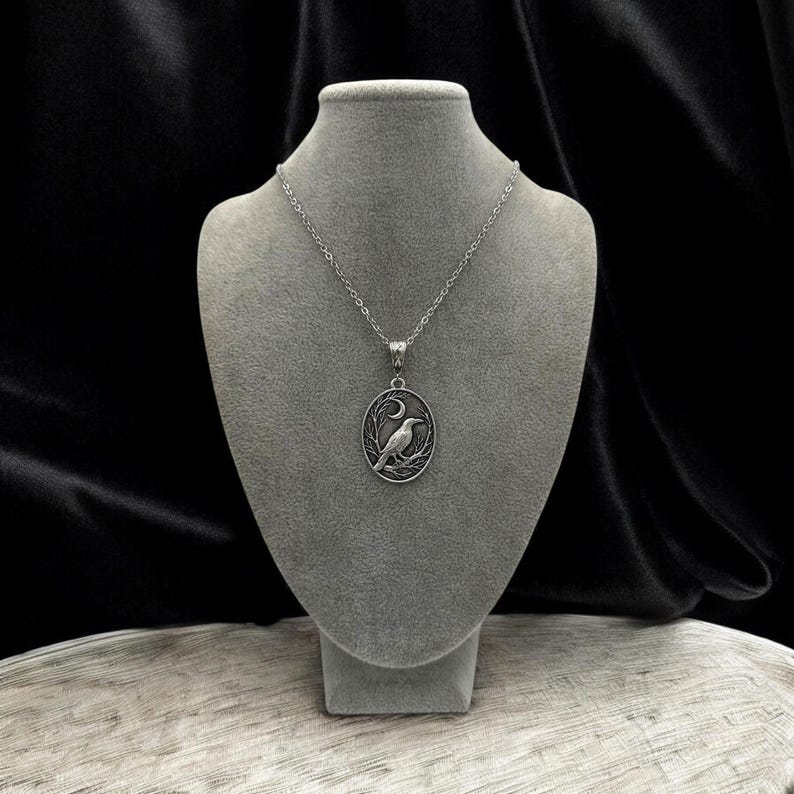 Puede incluir: Un collar con colgante ovalado de tono plateado que presenta un dise&ntilde;o en relieve de un cuervo posado sobre una rama bajo una luna creciente. El colgante cuelga de una delicada cadena plateada, mostrando detalles intrincados y un acabado met&aacute;lico.