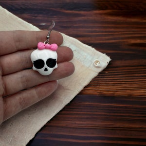 May include: White skull shaped earrings with a pink bow. The earrings are made of polymer clay and have a silver hook.