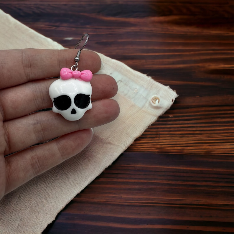May include: White skull shaped earrings with a pink bow. The earrings are made of polymer clay and have a silver hook.