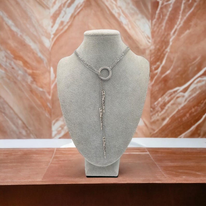 May include: A silver-toned lariat necklace featuring a circular pendant encrusted with small clear stones. Three delicate chains extend downward from the ring, each ending in a slender, textured, spike-shaped charm with a metallic finish.