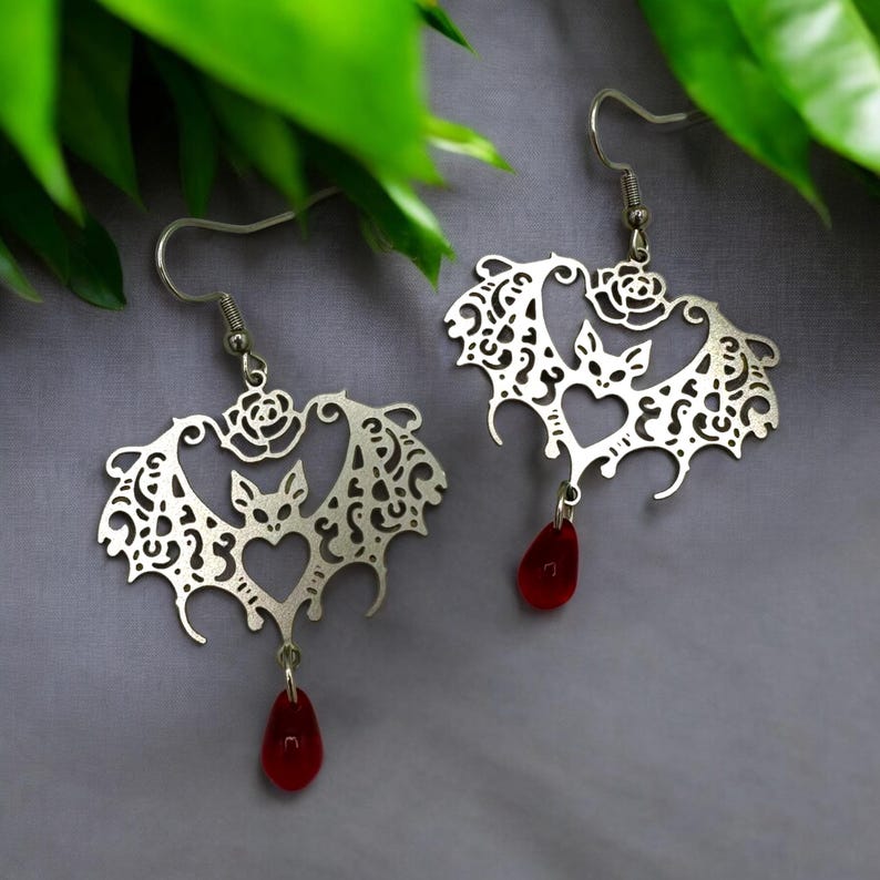 May include: A pair of silver earrings shaped like bats with intricate cutouts and red teardrop-shaped crystals hanging from the bottom.