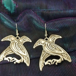 May include: A pair of silver earrings shaped like stylized ravens. The ravens are intricately detailed with swirling patterns and have a three-dimensional look.
