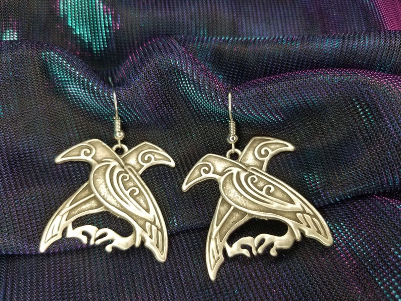 May include: A pair of silver earrings shaped like stylized ravens. The ravens are intricately detailed with swirling patterns and have a three-dimensional look.