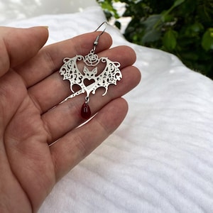 May include: Silver bat earring with a red teardrop pendant. The bat is detailed with a filigree design and a rose at the top.