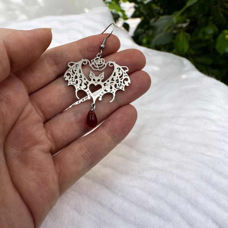 May include: Silver bat earring with a red teardrop pendant. The bat is detailed with a filigree design and a rose at the top.