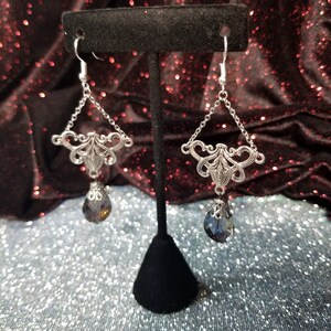 May include: Silver dangle earrings with a teardrop-shaped crystal pendant. The crystal is a dark blue color with a slight iridescent shimmer.
