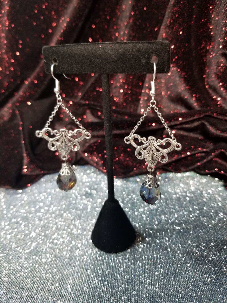May include: Silver dangle earrings with a teardrop-shaped crystal pendant. The crystal is a dark blue color with a slight iridescent shimmer.