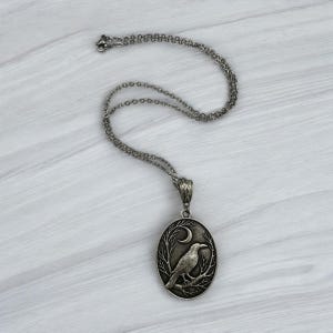 Puede incluir: Un collar con colgante de metal plateado que presenta un dije de forma ovalada. El colgante muestra un dise&ntilde;o en relieve de un p&aacute;jaro posado sobre una rama bajo una luna creciente. El dije cuelga de una delicada cadena de eslabones plateados.