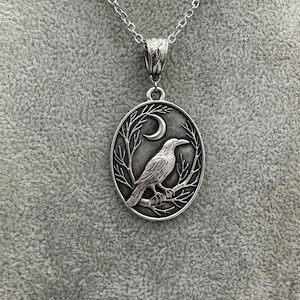 Puede incluir: Un collar con colgante ovalado de tono plateado que presenta un dise&ntilde;o en relieve de un cuervo posado sobre una rama de &aacute;rbol desnuda bajo una luna creciente. El colgante cuelga de una cadena de metal plateado con un enganche ornamentado.