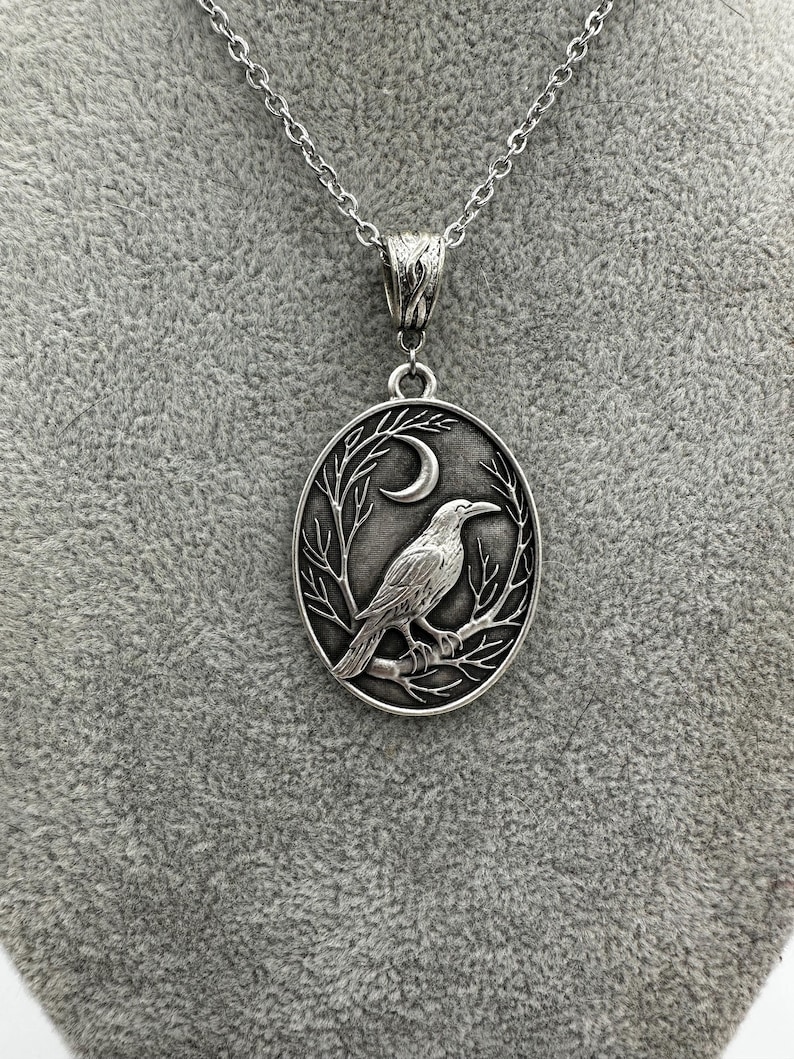 Puede incluir: Un collar con colgante ovalado de tono plateado que presenta un dise&ntilde;o en relieve de un cuervo posado sobre una rama de &aacute;rbol desnuda bajo una luna creciente. El colgante cuelga de una cadena de metal plateado con un enganche ornamentado.