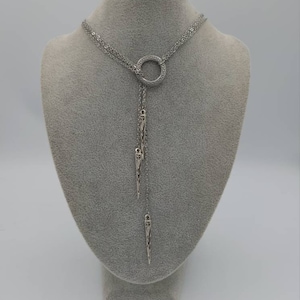 May include: A silver-toned lariat necklace featuring a central circular ring with a textured pattern. Three delicate chains descend from the ring, each ending in a slender, pointed metal pendant with intricate engraved detailing along the surface.