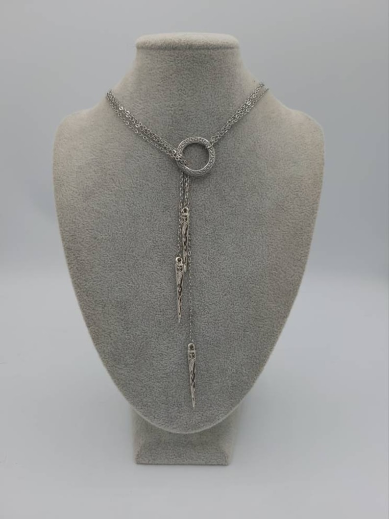 May include: A silver-toned lariat necklace featuring a central circular ring with a textured pattern. Three delicate chains descend from the ring, each ending in a slender, pointed metal pendant with intricate engraved detailing along the surface.