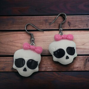 May include: A pair of white skull earrings with pink bows. The earrings are made of polymer clay and have a glossy finish.