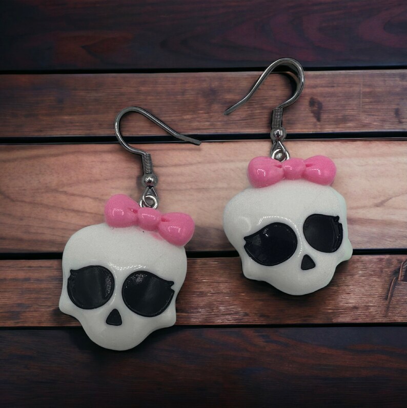 May include: A pair of white skull earrings with pink bows. The earrings are made of polymer clay and have a glossy finish.