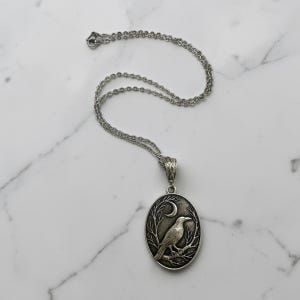 Puede incluir: Un collar con colgante ovalado de tono plateado que presenta un relieve detallado de un cuervo posado en una rama bajo una luna creciente. El colgante cuelga de una delicada cadena plateada con una anilla texturizada.