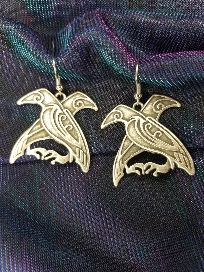 May include: A pair of silver earrings shaped like stylized ravens. The earrings have intricate details and a dangling design.