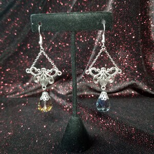 May include: A pair of silver dangle earrings with a teardrop-shaped crystal. The crystal on the left earring is a light amber color, while the crystal on the right earring is a dark blue color.