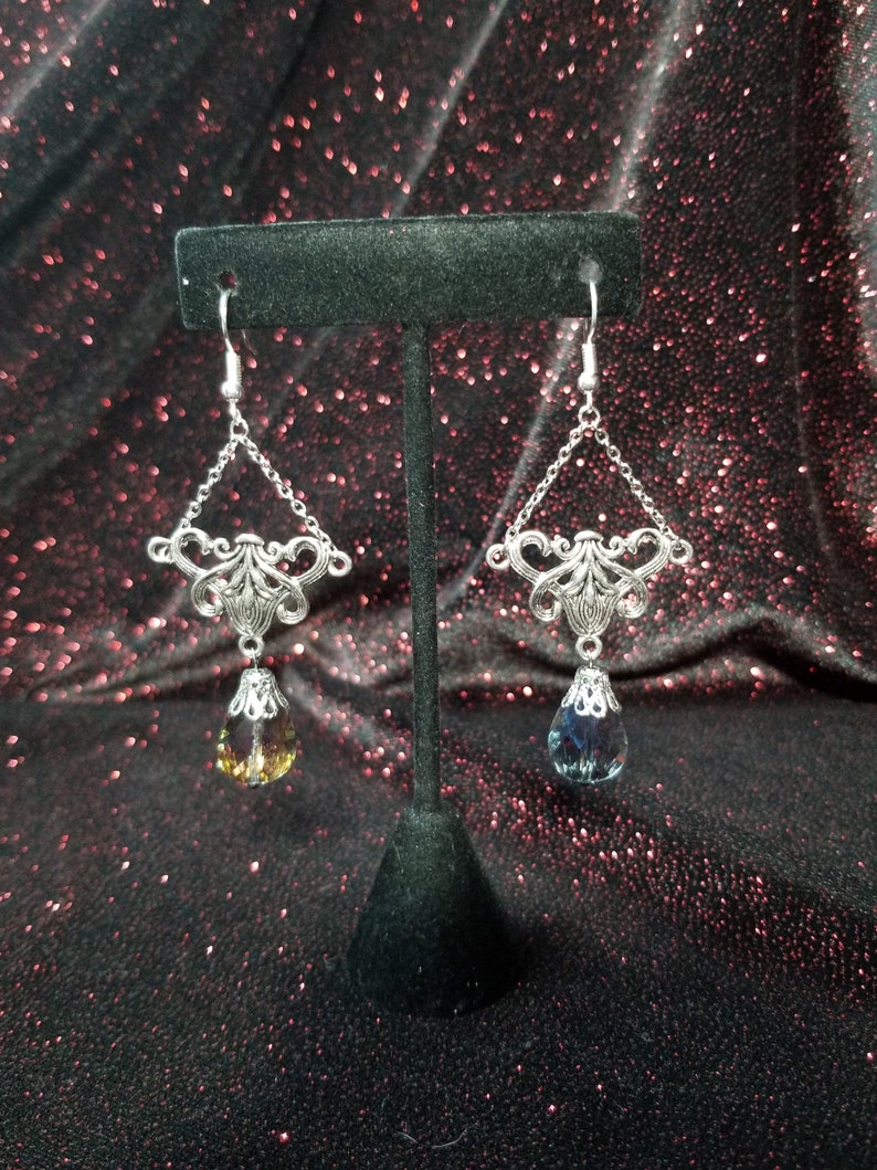 May include: A pair of silver dangle earrings with a teardrop-shaped crystal. The crystal on the left earring is a light amber color, while the crystal on the right earring is a dark blue color.