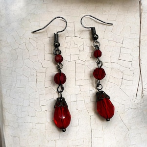 Peut inclure: Une paire de boucles d'oreilles pendantes avec des perles de verre rouges et des breloques en forme de goutte. Les boucles d'oreilles ont une finition en métal foncé.