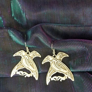 May include: A pair of silver earrings featuring a detailed design of two birds with intricate patterns. The earrings are hanging from a silver hook.