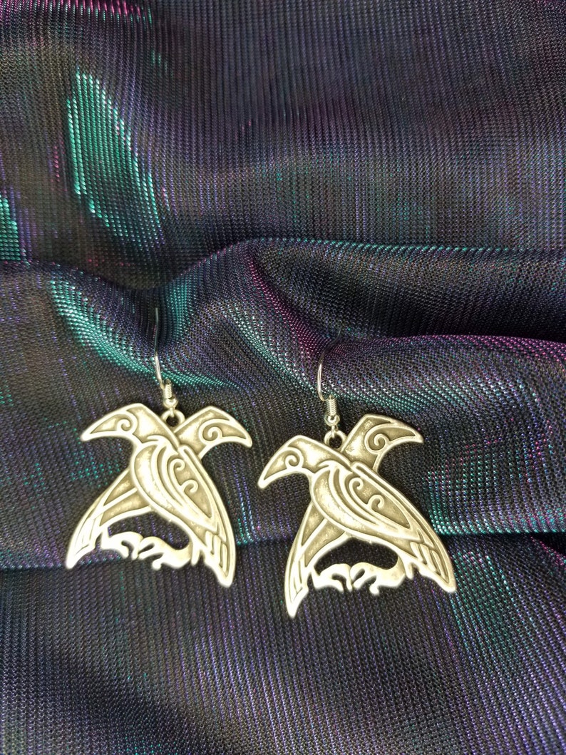 May include: A pair of silver earrings featuring a detailed design of two birds with intricate patterns. The earrings are hanging from a silver hook.