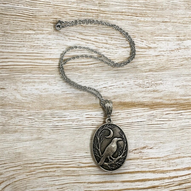 Puede incluir: Un collar con colgante ovalado de tono plateado que presenta un relieve de un cuervo posado en una rama bajo una luna creciente. El colgante cuelga de una delicada cadena plateada con una anilla texturizada que une ambas piezas.
