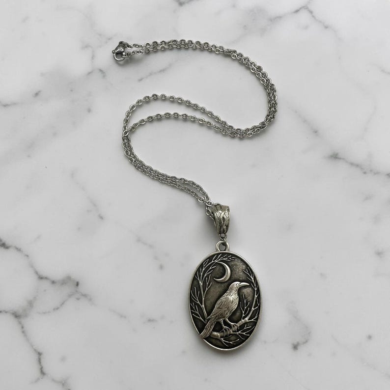 Puede incluir: Un collar con colgante ovalado de tono plateado que presenta un dise&ntilde;o en relieve detallado de un cuervo posado en una rama bajo una luna creciente. El colgante cuelga de una delicada cadena de metal plateado con un cierre seguro.