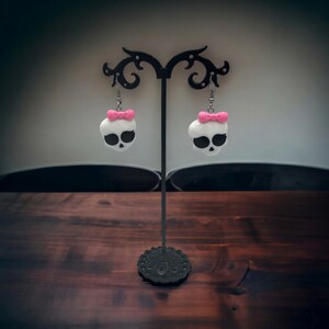 May include: A pair of white skull earrings with pink bows hanging on a black earring stand. The earrings are on a wooden surface.