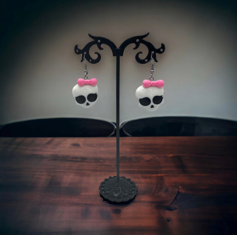 May include: A pair of white skull earrings with pink bows hanging on a black earring stand. The earrings are on a wooden surface.