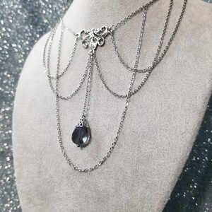 May include: A silver chain necklace with a delicate, ornate pendant and a single, dark blue gemstone hanging from the bottom.