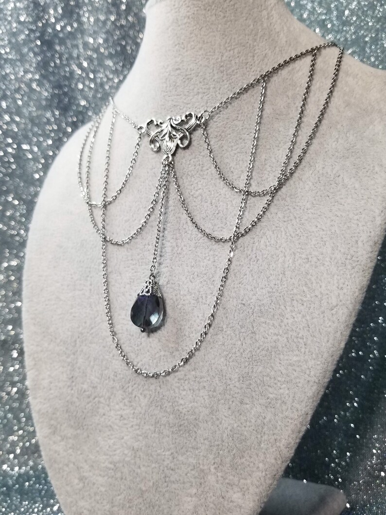 May include: A silver chain necklace with a delicate, ornate pendant and a single, dark blue gemstone hanging from the bottom.