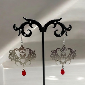 May include: Silver dangle earrings with a bat and rose design and a red teardrop crystal.