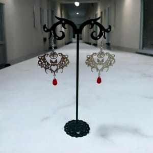 May include: A pair of silver earrings with a bat design and red crystal drops. The earrings are displayed on a black stand.