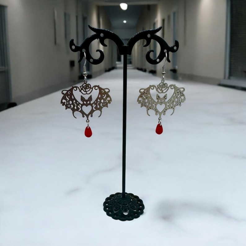 May include: A pair of silver earrings with a bat design and red crystal drops. The earrings are displayed on a black stand.