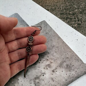 May include: A copper earring with a long, pointed, and slightly curved dangle. The dangle is attached to three copper beads with a spiral design.