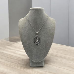 Puede incluir: Un collar de tono plateado con un colgante ovalado que presenta un dise&ntilde;o en relieve de un cuervo posado en una rama bajo una luna creciente. El colgante cuelga de una delicada cadena plateada, mostrando detalles intrincados y un acabado oscuro.