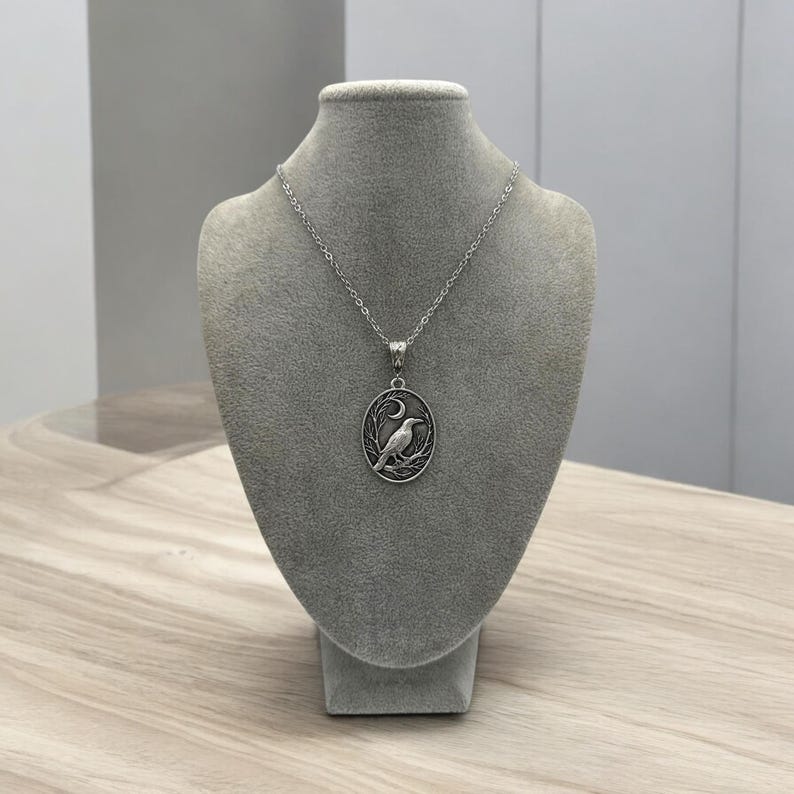Puede incluir: Un collar de tono plateado con un colgante ovalado que presenta un dise&ntilde;o en relieve de un cuervo posado en una rama bajo una luna creciente. El colgante cuelga de una delicada cadena plateada, mostrando detalles intrincados y un acabado oscuro.