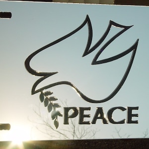 May include: A silver license plate with a black outline of a dove carrying an olive branch. The word "PEACE" is printed below the dove.