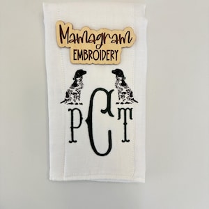 May include: White fabric with a monogram of the letters "PCT" in green thread. Two black and white dog silhouettes are flanking the monogram. A wooden sign with the text "Monogram Embroidery" is attached to the fabric.