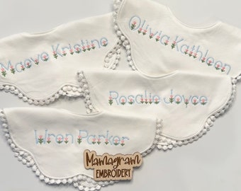May include: Four white cotton bibs with scalloped edges and embroidered names in blue with pink flowers. The names are Maeve Kristine, Olivia Kathleen, Rosalie Joyce, and Wren Parker. The bibs are arranged in a stack with the top bib visible and the bottom bib partially visible. A wooden sign with the text "Mamagram Embroidery" is in the bottom right corner.