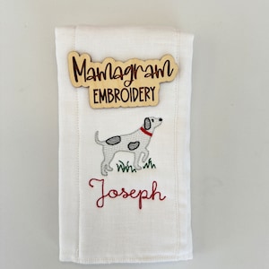 May include: White burp cloth with a black and white embroidered dog, green grass, and the name "Joseph" in red embroidery. A wooden sign with the text "Mamagram Embroidery" is attached to the cloth.