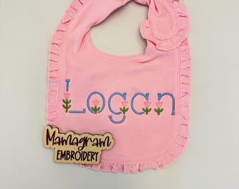 May include: Pink baby bib with a ruffled edge and the name "Logan" embroidered in blue with small pink flowers. A wooden tag with the text "Mamagram Embroidery" is attached to the bib.